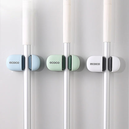 Ecoco mop and broom holders in pastel green and blue, mounted on a bathroom wall, securing cleaning tools with a sturdy grip.
